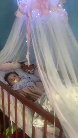 The video shows a baby lying in a crib under a sheer canopy adorned with fairy lights. The baby is wearing a blue and white gingham shirt and has a yellow pacifier in its mouth. The crib is filled with pillows, some of which have a grey and white grid pattern. A stuffed fox toy is visible in the crib. The overall scene is of a nursery, likely during the evening or nighttime due to the fairy lights. The baby appears calm and is looking upwards. The room has blue walls. There is some visible text on the pillows, including "SUPREME" and "HAVE SECRET". There is also some text on a label on a blanket at the bottom of the crib. The location is Tetiiv, Ukraine.