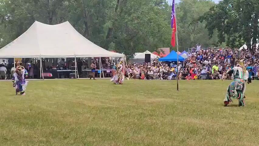 The video shows an outdoor event with a large white tent, a crowd of spectators, and people in traditional regalia dancing on a grassy field. In the foreground, three individuals are visible, two in elaborate regalia and one in a blue skirt and white top. The individual on the far left wears a blue and white feathered outfit. The individual on the right is dressed in a colorful outfit with intricate beadwork. In the middle ground, another person in regalia is performing a dance. Behind the dancers, a large white tent is set up, with people standing and sitting inside. Further back, a tiered seating area is filled with spectators. Two flags, one purple and one red, are planted on poles. The environment is a grassy field with trees in the background. The overall atmosphere appears to be a cultural celebration or powwow.