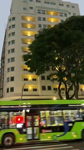 The video captures a street scene at dusk or night. A tall, modern building with numerous illuminated balconies dominates the background. In the foreground, a bright green bus is in motion, blurring due to its movement and the camera's perspective. The bus has its lights on, including red taillights. To the left of the bus, a traffic light is red, and a few pedestrians are visible on a crosswalk. A large, leafy tree stands to the right of the building, its branches partially obscuring the structure. The overall atmosphere suggests an urban environment with active street life.