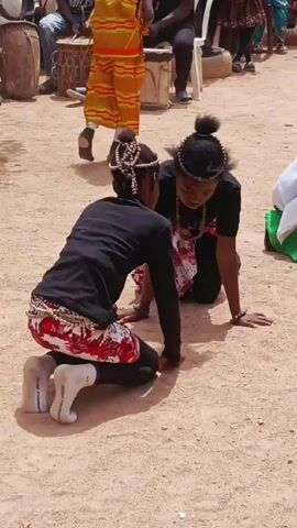 Several young women are seen performing a dance on the ground in an outdoor setting. The ground is covered with sand and there are other people and objects in the background. The dancers are dressed in black shirts and colorful leggings, with one wearing a green and white dress. They are moving their hands and bodies in a synchronized manner. Drums are audible in the background.