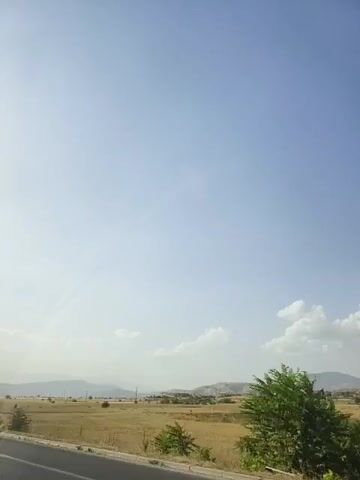 The video shows a vast, clear blue sky with a faint contrail stretching across it. Below the sky, there is a dry, golden landscape of fields with some scattered trees and distant hills. A paved road is visible in the foreground, along with a power line pole. The scene appears to be a rural area on a clear day.
