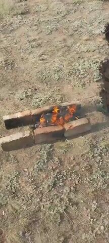 The video shows an outdoor scene with a small fire burning in a makeshift brick structure. The fire is in the center of the frame, surrounded by dry grass and dirt. To the right of the fire, there is a dirt embankment. To the left of the fire, there is a white plastic bag and a blue plastic bag filled with what appear to be pinecones or dried foliage. In the background, there is a metal samovar with a chimney, and a small table covered with items, including a pink and green cloth. The overall environment appears to be rural or undeveloped.