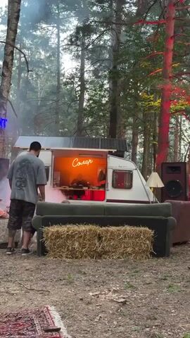 The video begins with a person standing with their back to the camera, facing a caravan parked in a wooded area. The caravan has solar panels on its roof and a neon sign that reads "Courage" in orange lettering above an open window. Inside the caravan, a person can be seen sitting down. A pinkish-purple smoke billows from the side of the caravan, near the person standing outside. In front of the caravan is a couch and a bale of hay. To the left of the caravan, a blue neon sign is partially visible. The ground is covered in dirt and leaves, with a red patterned rug laid out in the foreground. The trees in the background are tall and dense, with some red and yellow lights illuminating them from the right side of the frame. The overall atmosphere appears to be set up for an outdoor event or gathering.