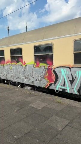 The video shows the side of a light yellow train car, heavily covered in graffiti. The graffiti is in various colors, including gray, black, red, and teal. The train car has several windows, some of which are reflecting the sky and trees. The train is parked on a gray tiled platform. The sky is blue with some white clouds. The graffiti obscures much of the train car's original paint and any text that might be present. The bottom portion of the train car is also covered in graffiti. There are some green plants growing between the tiles on the platform. The video pans from left to right, showing more of the graffiti. There is also some text on a dark blue section of the train car, which appears to say "Centralbahn - Sonder".