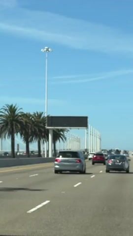 A dashcam view from a car traveling on a highway under a bright blue sky. Palm trees line the left side of the road, with a large overhead electronic sign displaying text. Several cars are visible on the highway ahead. The electronic sign transitions from a blank screen with a few red dots to displaying the message "ROADWO JCT 80 LANES C".