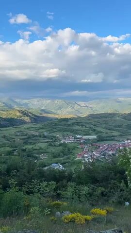 The video presents a wide-angle shot of a mountainous landscape beneath a partly cloudy sky. In the foreground, vibrant green vegetation is visible, including grass, small yellow flowers, and various trees. Below and slightly to the right, a small village is nestled amidst the green landscape. In the background, rolling hills stretch into the distance, layered with varying shades of green. The sky above is a bright blue, punctuated by scattered white clouds, some casting shadows on the land below.
