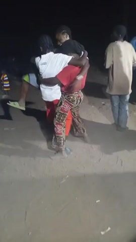 Here is a concise description of the video:

The video shows three people dancing closely in an outdoor setting, likely at night. One person is wearing a white shirt and red pants, another is wearing a red shirt and patterned pants, and the third is wearing green patterned shorts. They are all holding onto each other while moving, and the ground appears to be dirt or sand. Other people are standing in the background.
