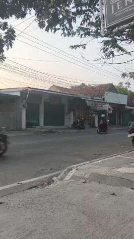The video captures a street scene with a gas station on the right. The sky is a pale orange, suggesting sunrise or sunset. Several shops with closed shutters are visible on the left side of the street. A few motorcycles are seen driving on the road. One motorcycle with two people is moving away from the camera. Another motorcycle is seen at the gas station. A person stands near the entrance of the gas station. The environment appears to be a typical urban or suburban street.
