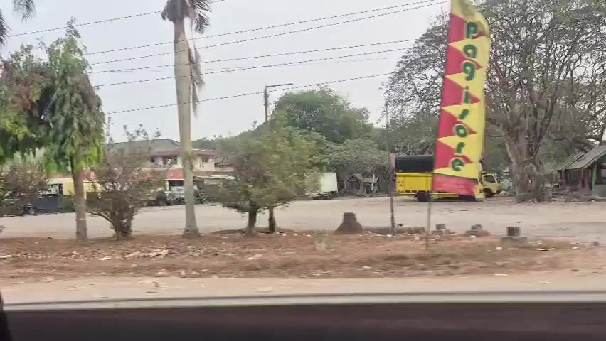 The video shows a view from a moving vehicle looking out at a street scene. In the foreground, there is a dusty, unpaved area with some scattered debris. Beyond this, a paved road is visible with several trees and utility poles. On the other side of the road, there are buildings, some with signs, and parked vehicles including trucks and cars. One building appears to be a restaurant or shop. Further in the distance, more trees and structures are visible. The sky is overcast. The camera appears to be panning or moving horizontally.