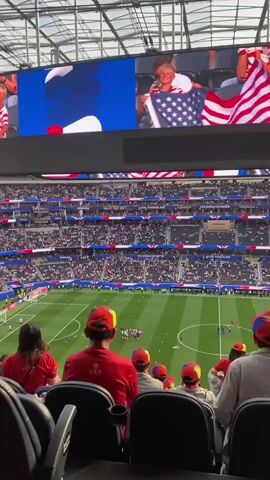 This video shows a packed stadium with a soccer match in progress. The focus is on the spectators, many of whom are wearing red and white, the colors of the American flag. A large screen at the top of the stadium displays images of people holding American flags and the word "USA" repeated multiple times. The crowd appears to be in a festive and patriotic mood, likely attending a sporting event where the United States is represented. Sunlight streams into the stadium, indicating it is daytime. The sign "349" is visible on an overhead structure, likely a seating section marker. The event seems to be a significant occasion, possibly a World Cup match, given the branding "FIFA WORLD CUP" visible on some stadium banners.