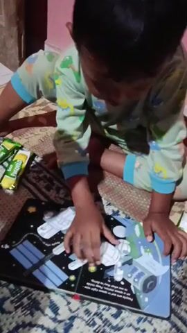 A young child wearing green pajamas with a dinosaur pattern is sitting on a patterned rug and looking at a pop-up book. The book is open to a page depicting a space scene with a space station and the Earth. The child is touching the pop-up elements of the space station with their right hand. There are two snack bars to the left of the book. The environment appears to be indoors, possibly a bedroom or living room.