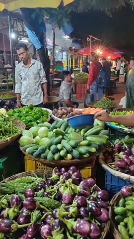The video shows a bustling outdoor vegetable market scene at night. The camera pans across stalls laden with fresh produce, including baskets of eggplants, cucumbers, gourds, cabbage, cauliflower, and green beans. Several vendors are visible, some interacting with customers or arranging their wares. The lighting appears to be a mix of overhead bulbs and natural ambient light, creating a somewhat dim and atmospheric setting. People are seen walking through the market, and the overall impression is one of a lively and active marketplace.