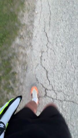 A man is walking down a cracked asphalt road as the sun sets in the background. He is wearing orange sneakers, black shorts, a black bandana, and mirrored sunglasses. He is shirtless and has a backpack slung over his shoulder. The landscape includes a field and some flowering bushes alongside the road.