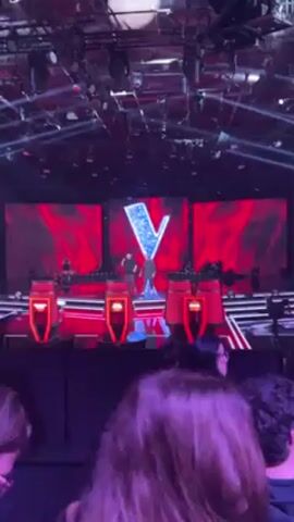 The video shows a stage set for "The Voice", with the signature large "V" logo displayed on a red screen at the back. The stage includes the four iconic red judge chairs. There are two men standing on the stage; one appears to be speaking into a microphone. A small crowd is seated in front of the stage. The lighting is bright with many spotlights illuminating the stage.