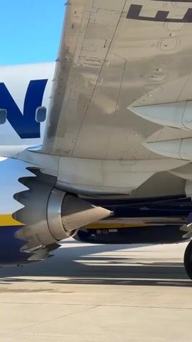 The video captures a Ryanair Boeing 737 MAX aircraft on the tarmac at Weeze Airport, Germany, during what appears to be a sunny day.

The initial frames offer a close-up of the aircraft's right wing and engine, highlighting the distinctive chevron-shaped exhaust nozzles characteristic of the 737 MAX series. The blue and yellow Ryanair livery, with the partial word "RYAN," is visible on the fuselage.

As the camera pans back, more of the aircraft's front section comes into view. Passengers are seen boarding the plane via a mobile stairway positioned at the forward door. Several individuals, including a person wearing a high-visibility vest, are on the tarmac. A piece of ground handling equipment, a small yellow vehicle, is partially visible in the background, along with a building that resembles an airport terminal.

The sky is clear and blue, indicating good weather conditions. Bright sunlight casts distinct shadows on the tarmac. The text "1G1" is visible on the side of the boarding stairs.