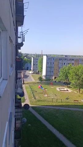 This is a vertical, eye-level outdoor shot of a residential area on a clear day. The camera is positioned on a balcony overlooking a courtyard with a playground. The foreground shows the side of a grey apartment building and a balcony with satellite dishes. In the mid-ground, there is a playground with a slide, swings, and a sandbox. Several trees are scattered throughout the courtyard, and pathways wind through the grassy areas. In the background, more apartment buildings and trees are visible under a bright blue sky. The overall impression is of a quiet, everyday scene in a housing complex.