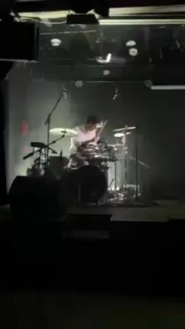 A low-quality recording shows a band performing on stage in a dimly lit club. The camera is positioned among the audience, focusing first on the drummer, wearing a white shirt, who is playing with energy. The camera then shifts to the keyboardist, dressed in dark clothing, who is also playing. The stage is dark, with lights shining from above. Audience silhouettes are visible in the foreground. The atmosphere suggests a live music performance in an intimate setting.