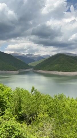 The video shows a lake surrounded by green hills and mountains under a cloudy sky. The water of the lake is calm and reflects the sky. The foreground shows lush green trees.