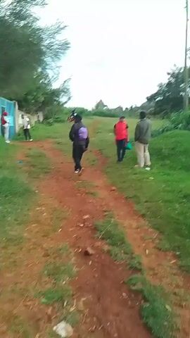The video shows a rural scene, likely in a developing country, with a red dirt path winding through green grass. Several people are seen walking along the path. The weather appears overcast with a cloudy sky. A wall made of concrete blocks is visible on the left side of the path. In the distance, there are trees and what appears to be a utility pole. Additionally, people can be seen standing near the wall and further up the path. A motorcycle passes by on the path, farther away from the camera.