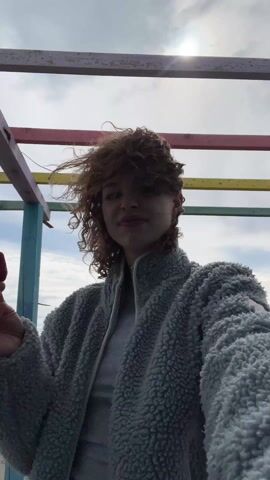 A person with curly red hair is shown in an eye-level shot, biting into a strawberry. They are wearing a grey fleece jacket and standing outside under a wooden structure painted with various colors, with the sky and ocean visible in the background. The person bites off part of the strawberry and looks at the camera before turning their head to the right. The sky is bright and the sun is shining.