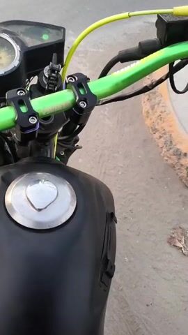 The video shows the view over the handlebars of a stationary motorcycle. The motorcycle has a black gas tank and a chrome gas cap. The handlebars are bright green with black hardware. The motorcycle's speedometer is visible. The motorcycle is parked on a paved surface next to a curb. The engine of the motorcycle is audibly idling.