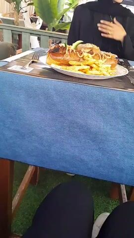The video shows a close-up view of a large hot dog served with a side of french fries on a white plate. The hot dog is in a bun and topped with what appears to be chili, sauce, and a cucumber slice. The fries are seasoned and piled around the hot dog. The plate is on a table covered with a blue cloth. In the background, there are hints of people, outdoor seating, and greenery, suggesting an outdoor dining setting. The camera angle is from above, looking down at the food.