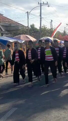 A group of young males wearing black caps, black jackets, and red and white striped shirts are walking down a street. Some of the individuals have fake beards and white face paint. One individual has a green bib with the number 069 on it. They are walking in what appears to be a parade or procession, as there are tents and stalls set up along the side of the road. The weather appears to be sunny.