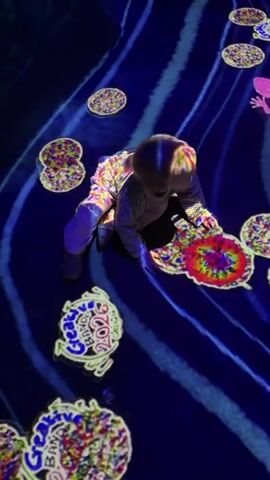 A young child with blonde hair wearing a striped long-sleeved shirt and light-colored pants is crawling on a dark blue surface. The surface is illuminated with projected images. There are wavy blue lines creating a water-like effect and numerous circular, colorful, abstract patterns spread across the floor. Some of these circular patterns have a yellow outline. In the background, a pink, stylized figure, possibly a mermaid or a human, and a yellow, lizard-like creature are also projected. The child is looking towards the right side of the frame and appears to be interacting with the projected images. The lighting is dim, with the projected images being the primary source of illumination.