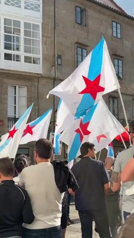 A group of people are gathered outdoors, holding several white flags with blue diagonal stripes and red stars. The flags are the Galician nationalist flag. The people appear to be standing on a street or plaza, with buildings visible in the background. The buildings are made of stone and have windows. The sky is not visible in the frame. The scene appears to be a demonstration or gathering.