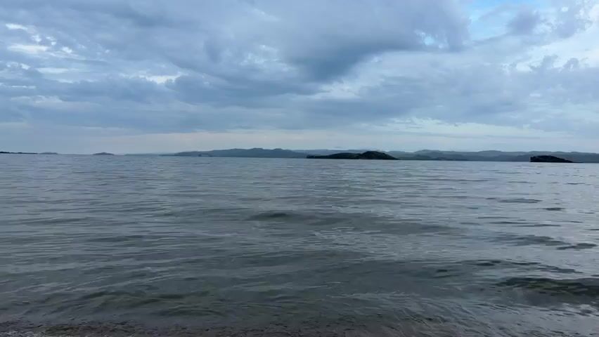 The video shows a calm body of water with small waves lapping at a sandy shore. In the distance, there are rolling hills or mountains and some small islands. The sky is overcast with dark clouds. The lighting suggests it is either early morning or late evening. There are no people or objects visible in the video besides the natural landscape.