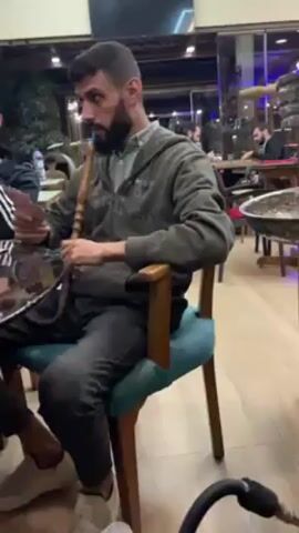A man with a beard is sitting in a chair, holding playing cards in his left hand and a hookah hose in his right hand. He is exhaling smoke from the hookah. He is wearing a green hooded sweatshirt and dark jeans.  The chair is wooden with a blue cushioned seat. In front of him is a low glass-topped table.  Another person is partially visible to his left, wearing an Adidas-striped shirt and tan shoes. The setting appears to be a restaurant or cafe, with other people seated at tables in the background. The floor is light-colored wood. There are plants visible behind the man through a glass window.