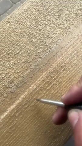 A hand is using a pointed tool to scratch letters into a wooden plank. The letters appear to be F, O, K. The plank has a rough texture and appears to be unfinished wood. The background is blurred but suggests an outdoor setting with paving stones visible.