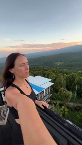 A person is taking a selfie from a balcony at sunset. The person is a light-skinned woman with long brown hair wearing a black athletic top and leggings. She smiles, raises an arm in the air, and turns the camera to reveal a view of mountains and a forest. The landscape is bathed in the golden light of the setting sun. In the foreground is a balcony with a dark railing. A white roof of a nearby building is also visible. The sky is a gradient of blue and orange with a few scattered clouds.