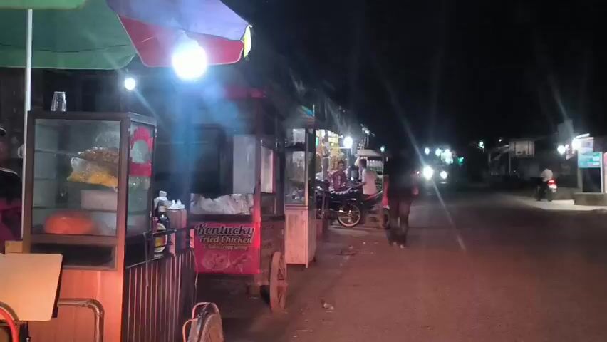 The video depicts a dimly lit street at night, possibly in a suburban or semi-urban area. Several food vendors with pushcarts are visible on the left side of the road, illuminated by individual lights. A man walks down the street away from the camera. Parked motorcycles line the side of the street further down. There are businesses across the street. Headlights from passing vehicles cast streaks of light. Several bags are piled on the side of the road near the businesses.