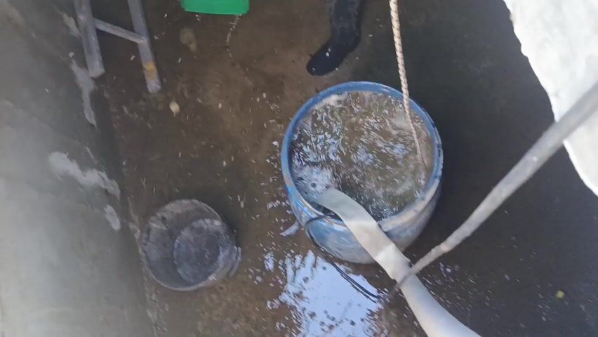 A man is seen in a concrete structure. The man is wearing black boots and a black shirt. He is holding a green bucket with a red lid and pouring water from the bucket into a blue barrel that contains water and a hose. A rope hangs down into the barrel. A small stepladder is leaning against one of the concrete walls. A second, gray bucket sits on the ground.