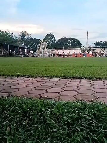 The video captures three individuals jogging along a paved pathway in an outdoor park or recreational area. The pathway is constructed from hexagonal bricks and is flanked by green grass. In the background, numerous trees are visible, along with distant structures including what appears to be a building with a traditional roof, and several tents or stalls. A tall, slender tower or antenna is also visible in the distance on the right side of the frame initially. The sky is bright with scattered clouds, suggesting a late afternoon or early evening setting. The individuals, dressed in athletic attire, run from left to right. One wears a grey shirt and black shorts, another a black and white patterned shirt and black shorts, and the third a red shirt and black shorts. The scene depicts a tranquil environment suitable for exercise, located in Matesih, Indonesia.