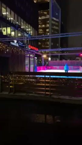 The video depicts a small, open-air ice rink in an urban setting at night. Several people are ice skating. The rink is surrounded by a wooden barrier decorated with strings of lights. Overhead, there is a metal framework with colorful stage lighting in shades of blue, green, and pink.  Tall buildings with illuminated windows surround the rink. The ground in the foreground is wet, reflecting the lights. A sign with the word "БАНКИРЫ" in Cyrillic is visible on a building adjacent to the rink.