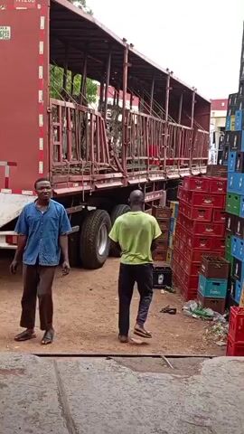 Two men stand in a dirt lot next to a large red trailer truck. The truck's rear section is open, revealing a metal cage-like structure. On the right side of the scene, stacks of red and green plastic crates are piled high. The man on the left wears a blue short-sleeved shirt and brown pants. The man on the right wears a lime green t-shirt and dark pants. The man on the right appears to be walking away from the truck. The ground is covered in dirt and some debris. The lighting suggests it is daytime.
