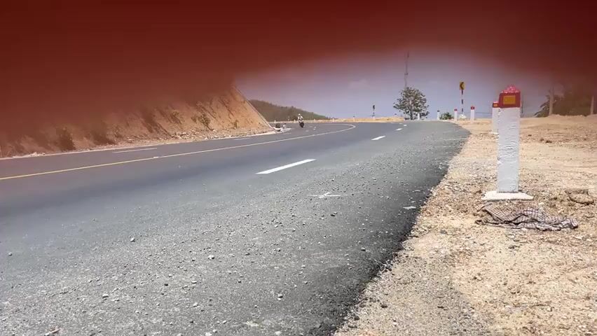 The video shows a lone motorcyclist riding on an asphalt road. The road curves gently and is marked with yellow and white lines. The motorcycle is moving from left to right. A grassy bank borders the left side of the road, while the right side features a sandy area with a white post marked in red. In the distance, a tree and several other posts are visible. The scene is bathed in bright sunlight. A blurred orange or red color obscures the upper section of the video.
