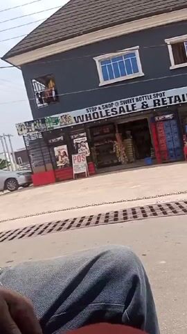 From a moving vehicle, the video captures a street scene with commercial buildings. The first building is dark gray with "STOP & SHOP @ SWEET BOTTLE SPOT WHOLESALE & RETAIL" signage and displays stacks of crates and bottles. The adjacent buildings are a mix of gray and dark gray, with signs for "POS PHONE SMART ACCESSORIES" and "GOODNEWS COSMETICS" along with a food store with a pink umbrella and menu board. Mannequins stand in front of the buildings. Power lines are visible overhead. A chain is strung across the street. The viewer is seated in the vehicle, wearing blue jeans.