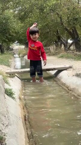 A young boy wearing a red long-sleeved shirt and dark shorts stands in a shallow irrigation ditch filled with murky water. He is looking to his left and smiling, with his right arm raised. The ditch has concrete sides and a narrow wooden plank crosses it a short distance ahead of the boy. Trees with green foliage line the sides of the ditch, and dry ground is visible beyond them. The boy splashes in the water and moves his hands through it.