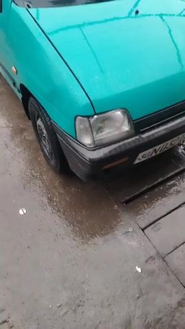 The video shows a light turquoise-colored car in what appears to be an outdoor car wash area. The car is wet, suggesting it has recently been washed or it is raining. The camera pans slightly from the front of the car to reveal more of the surrounding environment. In the background, there are other cars parked under a metal framework, also appearing wet. The ground is concrete and is also wet. A person can be seen standing near the other cars, possibly involved in washing or detailing them. The overall atmosphere seems damp and utilitarian, focusing on the car wash environment.