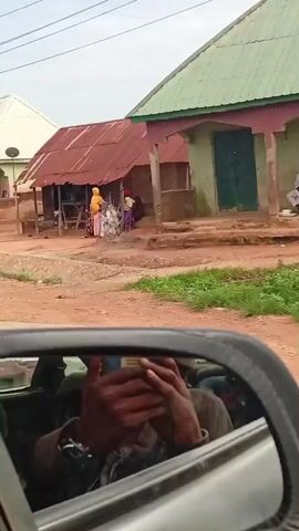A brief video captures a street scene in what appears to be a rural or semi-urban environment. The footage is filmed from inside a vehicle, with a side mirror prominently in the foreground, reflecting the interior of the car and a person covering their face.

The street is unpaved and dusty. On the left side of the street, there are buildings with corrugated metal roofs and pink-painted walls, some appearing to be shops or residences. In front of one building, a stack of tires is visible. Further down the street, another building has a white roof.

Several people and vehicles are seen in the video:
- A man in a light blue shirt and dark pants walks with his hands behind his head.
- Two young boys are seen running in the distance.
- Two men ride a bicycle, one pedaling and the other sitting behind him.
- Two men ride a motorcycle. One is wearing a red hooded sweatshirt and the other is wearing a dark blue cap.

The overall impression is a snapshot of daily life in a community, with people moving about and engaging in various activities. The presence of the vehicle suggests movement through the area.