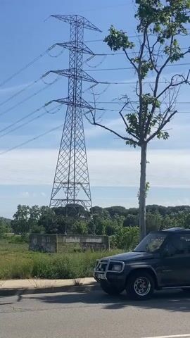 The video opens with a dark gray two-door SUV, identified by the text "Vitara" on its side, parked on the edge of a paved road. To the right of the vehicle, a tree with green leaves stands. In the background, a tall, prominent metal lattice electricity pylon is visible, with multiple power lines stretching across a clear blue sky dotted with a few wispy white clouds. Beyond the road, the landscape consists of green grassy areas and distant trees.

The camera then slowly pans from left to right, revealing more of the surroundings. The large electricity pylon remains in view, with its power lines extending into the distance. As the pan continues, another, smaller pylon can be seen further away, connecting to the network of power lines. The background features rolling green fields and densely forested mountains or hills under the bright, sunny sky. The foreground continuously shows the paved road, a concrete curb, and a strip of green grass.

The scene depicts an open, rural or suburban landscape, captured on a clear and sunny day. There are no people visible, and the only discernible action is the smooth, deliberate panning of the camera across the serene environment in Santa Perpètua de Mogoda, Spain.
