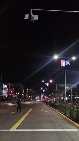 The video captures a nighttime scene in a city. The camera faces down a street, showing streetlights, traffic signals, and illuminated flags lining the roadway. A person walks down the street on the left. Traffic is sparse.  A sign indicates "Corredor Buses" and "Otros Vehiculos". The sky is dark, and the buildings on either side of the street are dimly lit. The overall atmosphere suggests a quiet urban setting at night.