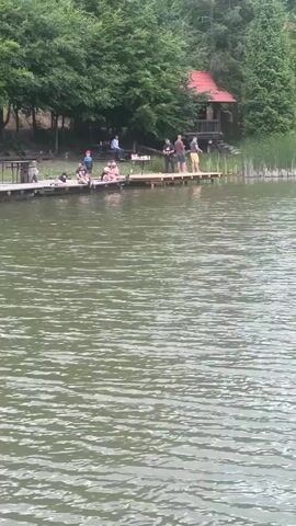 A group of people are gathered on a wooden dock by a lake. Some are sitting on the dock, while others are standing. One person on the dock appears to be fishing. In the background, there is a red-roofed wooden cabin nestled amongst trees. The water is calm with ripples on the surface. The overall atmosphere appears to be relaxed and casual, possibly a recreational outing.