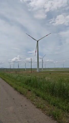 The video shows a roadside view of a wind farm on a partly cloudy day. The camera appears to be mounted inside a moving vehicle, facing the right side of the road where the wind turbines are located. There is a paved road with a patch of green grass alongside it. Beyond the grass is a field with several wind turbines that are actively rotating. Power lines and utility poles are visible in the background. The sky is filled with clouds, some of which are puffy and white. There is no evidence of people in the video.