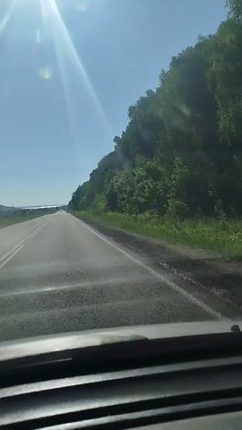 The video shows a view from the driver's seat of a car traveling on a paved road. The road has two lanes, with solid white lines marking the edges and dashed white lines separating the lanes. The sun is shining brightly in the clear blue sky, casting glare on the windshield. To the right of the road, there is a densely wooded hillside. To the left of the road, there are fields and a body of water in the distance. The car appears to be moving at a moderate speed.