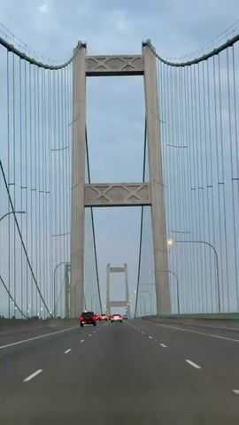 The video shows a first-person perspective of driving across a large suspension bridge. The camera is positioned within a vehicle, looking forward through the windshield. The bridge has massive concrete towers with vertical cables extending from them, forming the suspension system. The roadway is paved and has lane markings. Several vehicles are visible ahead on the bridge, traveling in the same direction. The sky is overcast with a pale, diffused light, suggesting it might be dawn, dusk, or a heavily clouded day. The overall impression is of a journey across a significant piece of infrastructure.