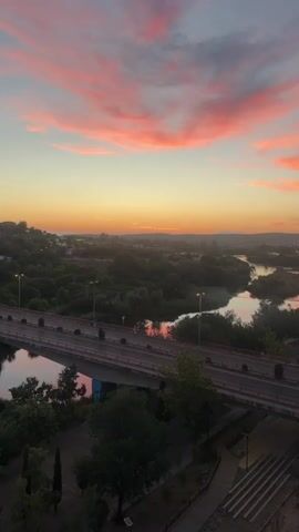 A vertical video shows a picturesque sunset over a river and a bridge. The sky is a canvas of warm oranges and pinks, with wispy clouds catching the last rays of the sun. A winding river flows through a lush green landscape, dotted with trees and buildings. A bridge spans the river, carrying traffic across. The foreground shows a pathway leading down a set of stairs, with trees and plants adding to the natural beauty of the scene. A lone car drives across the bridge, its headlights illuminating the road ahead. The video captures the serene and tranquil atmosphere of a sunset over a peaceful landscape.